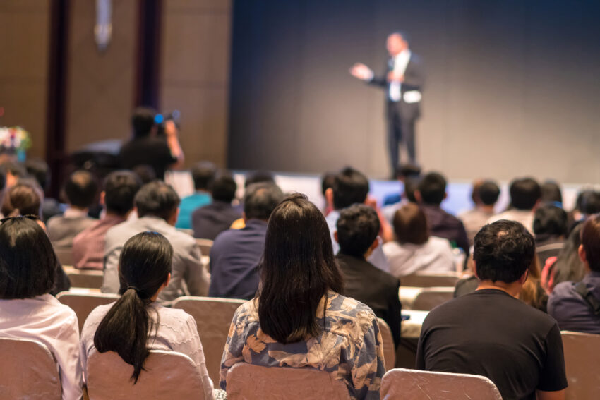 Rear side of Audiences sitting and listening the speackers on the stage in low light conference hall, event and seminar concept